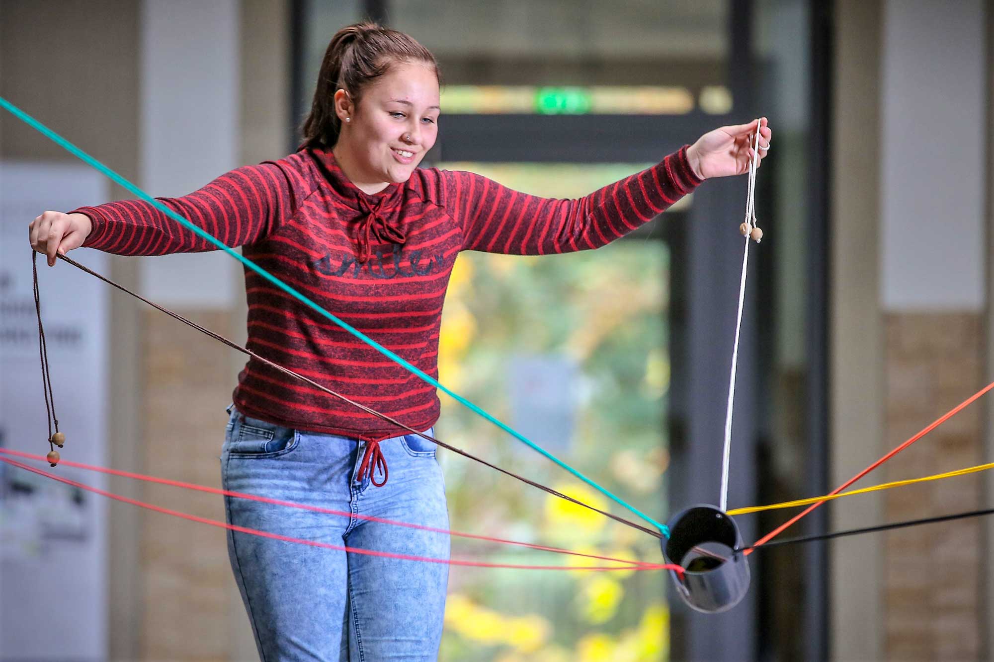 Eine Schülerin spielt ein Gruppenspiel. Sie hält zwei Seile fest, die an einem kleinen Rohr befestigt sind. Von dem Rohr gehen noch 5 weitere Seile ab und Enden außerhalb des Bildausschnittes.