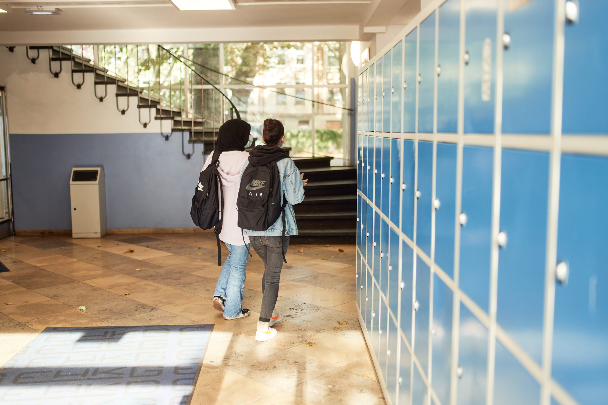 Schulflur, zwei Schulkinder laufen auf eine Treppe zu. Rechts sind blaue Schließfächer zu sehen.